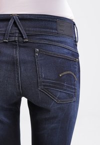 Dunkelblaue Slim-Fit-Jeans aus Denim, mit einem Markenpatch, Gesäßtaschen mit gelben Nähten und einer leicht verblassten Textur.