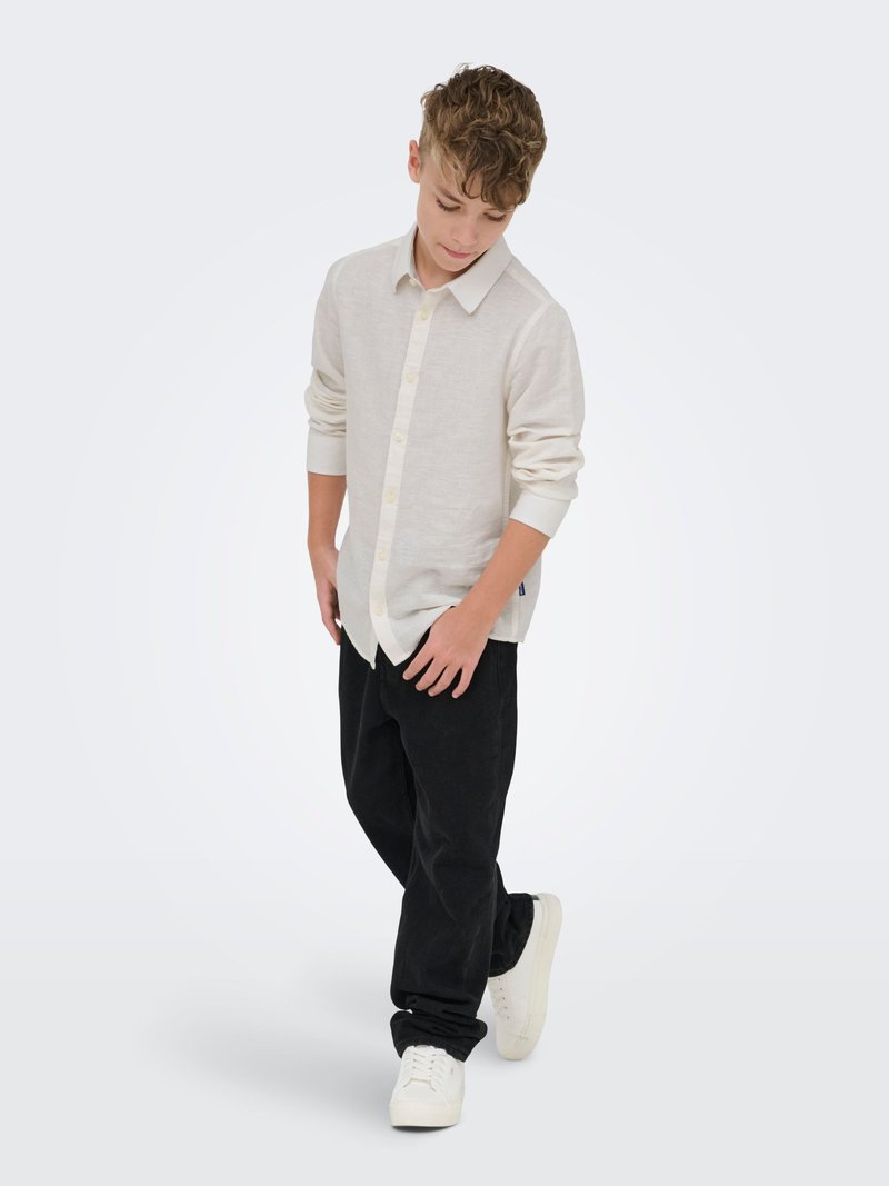 Garçon aux cheveux bouclés portant une chemise beige clair à boutons, un pantalon noir et des baskets blanches, regardant vers le bas avec un pied légèrement levé.