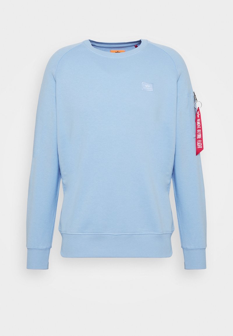 Sweat-shirt bleu clair à manches longues avec col ras du cou, poignets et ourlet côtelés, petit logo sur la poitrine et étiquette rouge "Remove Before Flight" sur la poche à fermeture éclair de la manche gauche.