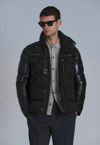 Zwarte puffy jas met glanzende panelen, hoge kraag en twee borstzakken, gecombineerd met een lichtgrijs overhemd met knopen en donkere broek.