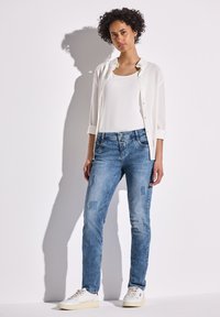 Jeans de mezclilla azul con parches desgastados, combinados con una camiseta sin mangas blanca y una camisa ligera de botones. Zapatillas blancas completan el look.