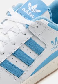 Vita och blå Adidas-sneakers med tre strukturerade blå ränder, vita snörningar och Adidas-logotyp på tungan och sidopanelen.