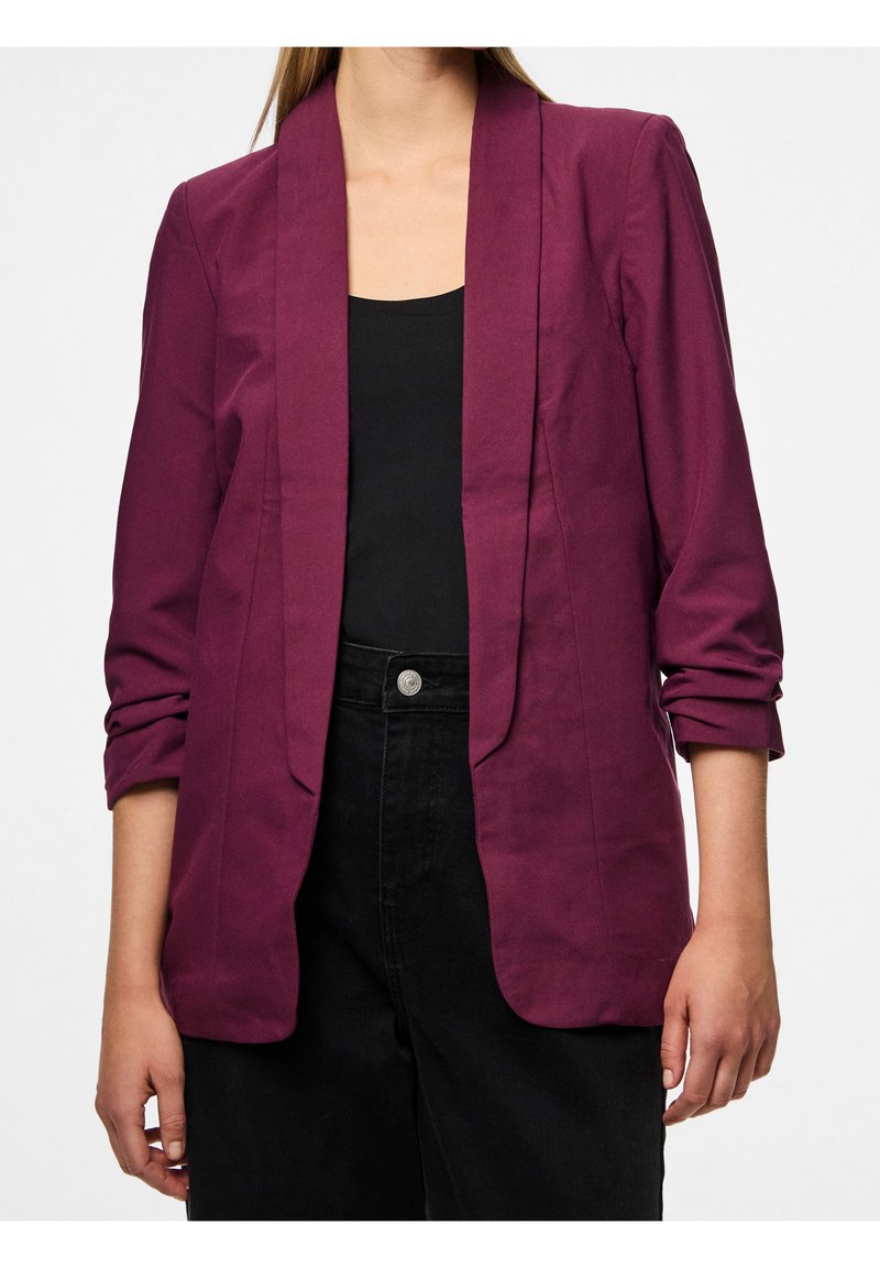 Femme portant un blazer violet foncé avec les manches retroussées sur un haut noir et un pantalon noir taille haute, debout devant un fond uni.