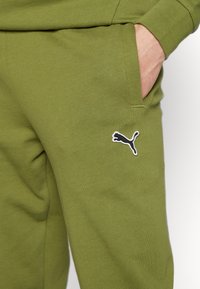 Gröna bomullssweatpants med en sidoficka och en svart logga på låret. Texturen är slät med en avslappnad passform.