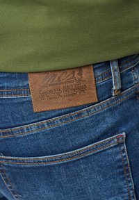 Une pièce en cuir marron avec du texte en relief sur un jean en denim, comportant un tissu bleu, des coutures orange et un tissu texturé vert au-dessus de la ceinture.