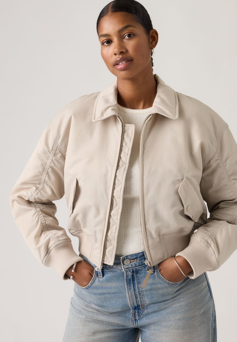 Femme les mains dans les poches portant une veste bomber beige clair sur un haut en maille blanc et un jean en denim bleu, debout devant un fond uni.