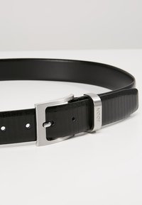 Schwarzer Ledergürtel mit strukturiertem Finish, ausgestattet mit einer silbernen Metallschnalle und einer Logo-Akzentuierung in der Nähe der Schnalle. Verstellbar mit mehreren Löchern.