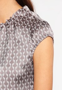 Blouse grise en tissu à motif texturé avec des demi-cercles blancs. Les manches sont froncées à l'épaule, créant une silhouette douce.
