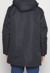 Cappotto blu navale con cappuccio, dalla finitura testurizzata, maniche lunghe, orlo che si estende sotto la vita e dettagli di design minimalisti.