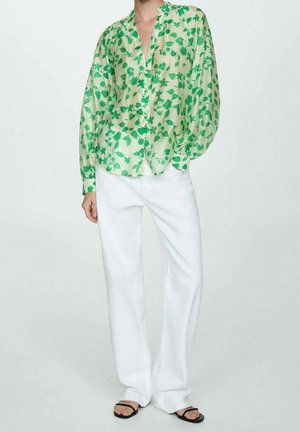 Personne portant un chemisier fleuri vert clair avec de longues manches bouffantes, un pantalon blanc à jambes larges et des sandales noires à bouts ouverts, debout devant un fond uni.