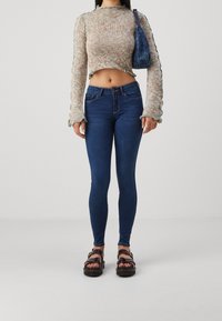 Suéter de punto multicolor, cortado, con mangas acampanadas largas, combinado con jeans ajustados azul oscuro y sandalias negras con tiras. El bolso tiene un diseño texturizado.