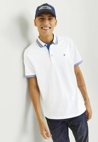 Weißes Poloshirt mit blauer Kragen- und Besatzfarbe, mit zwei Knöpfen, aus strukturiertem Stoff, getragen mit dunkelblauen Jeans. Blauer Hut mit Text oben.