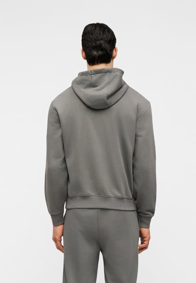 Homme portant un sweat à capuche gris uni et un pantalon gris assorti, debout de dos sur un fond clair uni.
