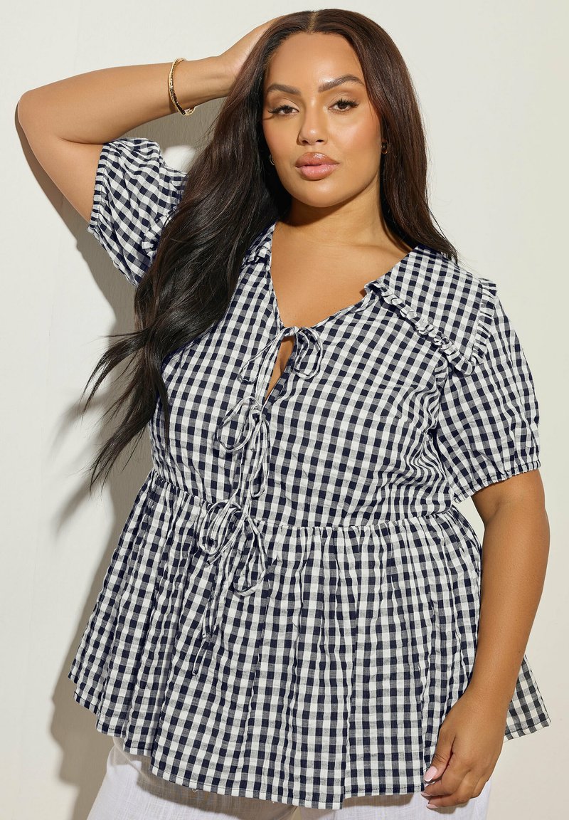 Kvinde med langt mørkt hår iført en sort-hvid ternet bluse med bindebånd, poserer med den ene hånd på hovedet mod en ensfarvet baggrund.