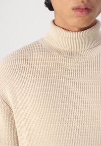 Pull à col roulé beige en tricot avec un motif texturé, doté d'un col côtelé et d'une coupe légèrement ample.