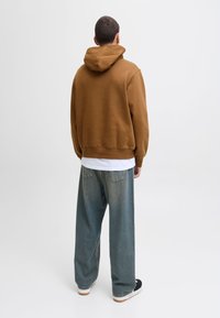 Brązowa bluza z kapturem o luźnym kroju, noszona na białej koszuli, zestawiona z luźnymi jasnoniebieskimi dżinsami i czarnymi butami.