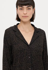 Camisa de manga longa em padrão de leopardo nas cores castanho escuro e preto, com colarinho tipo notched, bolso frontal e fecho com botões. Textura de tecido suave.