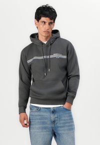 Hoodie gris avec un cordon de serrage, rayures horizontales blanches et bleues, poche avant, poignets et ourlet côtelés. Porté sur un jean bleu clair.