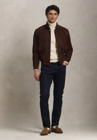 Rjav suedni jopič, kremni pulover na vratu, temne kavbojke in rjave mokasinke. Model stoji ob nevtralni ozadju in prikazuje eleganten, sodoben slog.