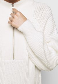 Pull en tricot blanc avec un col montant et une demi-fermeture éclair, présentant une texture côtelée et des épaules sans couture. Tissu doux et épais avec une coupe décontractée.