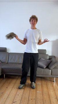 Modèle en t-shirt blanc et pantalon noir, tenant un chapeau gris, se tenant sur un sol en bois près d'un canapé en cuir gris avec des coussins.