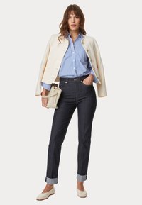 Chaqueta blanca texturizada sobre una camisa de botones a rayas azul y blanca. Jeans de mezclilla oscura con los puños doblados y zapatillas planas de color crema.