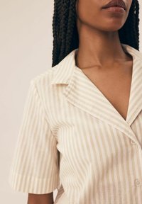 Jasnobeżowa koszula z pionowymi białymi paskami, z kołnierzykiem i krótkimi rękawami. Posiada zapięcie na guziki i gładką teksturę.