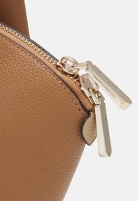 Róg torby z teksturowanej skóry w odcieniu brązowym z podwójnymi, złotymi zamkami błyskawicznymi z wygrawerowanym napisem "kate spade" na beżowej taśmie zamka.