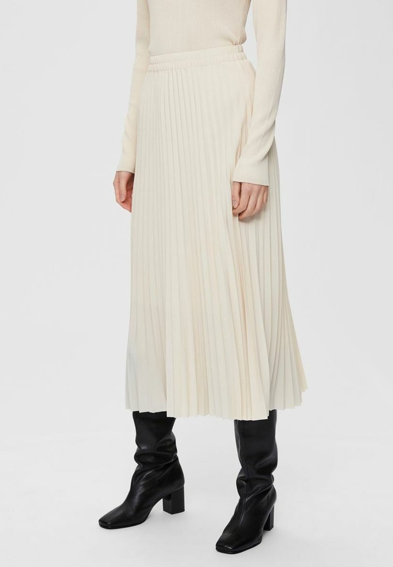 Gonna midi plissettata in crema, con una vita elasticizzata e un design fluido. Abbinata a stivali neri aderenti al ginocchio.