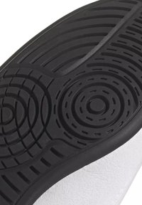 Semelle en caoutchouc noir avec des motifs de bande de roulement circulaires et linéaires sous une chaussure blanche.