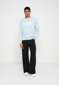 Gaiši zils sporta krekls ar baltu logotipu, kombinēts ar melnām platām džinsa biksēm un baltām kedām. Gluda auduma tekstūra un brīvs piegriezums.