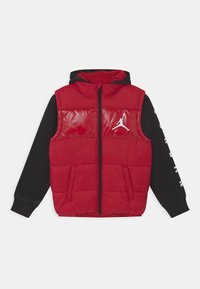 Czerwona pikowana kamizelka o błyszczącej fakturze, czarne długie rękawy, przedni zamek błyskawiczny, dwie boczne kieszenie oraz biały logo na piersi. Projekt z kapturem.