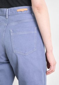 Calças de ganga lilás claras com corte reto, apresentando um único bolso na parte de trás e uma etiqueta de couro na cintura.