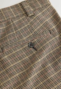 Pruun ja must houndstooth mustriga kangas, millel on nööbi sulgemine ja voodriga taskud, millel on tekstureeritud viimistlus.