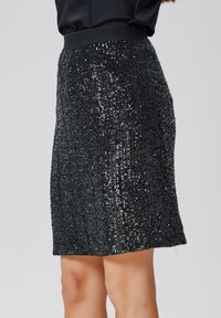 Jupe en sequins noirs avec une taille ajustée, présentant une surface texturée et des détails scintillants. Longueur mi-cuisse et silhouette droite.