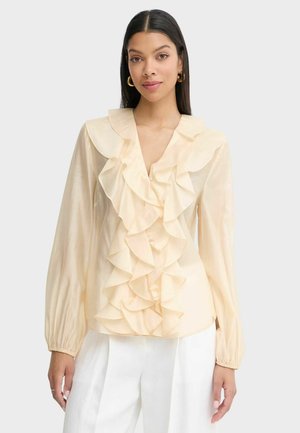 Donna che indossa una camicetta color crema a maniche lunghe con grandi ruches sul davanti e pantaloni bianchi, posando su uno sfondo chiaro e uniforme.