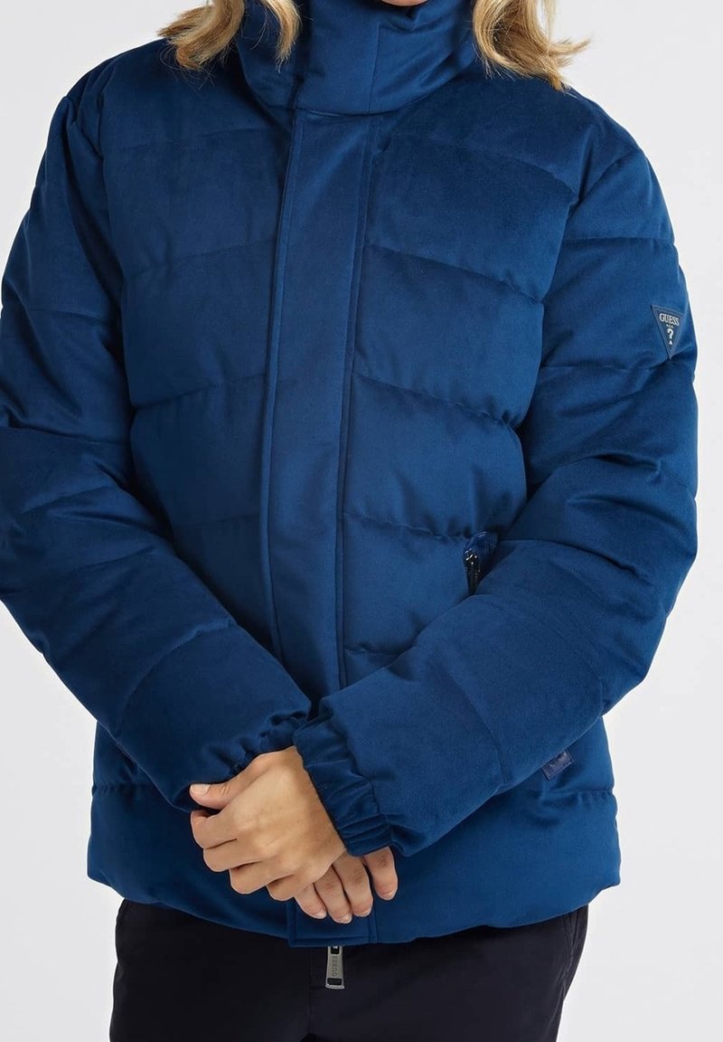 Femme portant une doudoune matelassée bleue avec des poignets élastiques et un patch du logo de la marque sur la manche gauche, les mains jointes devant elle.