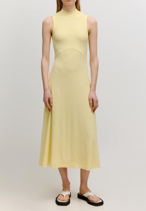 Ärmelloses, enganliegendes Kleid aus hellgelbem Rippstoff. Bodenlang mit einem hohen Kragen, betonter Taille und fließendem Rock. Kombiniert mit flachen Sandalen.