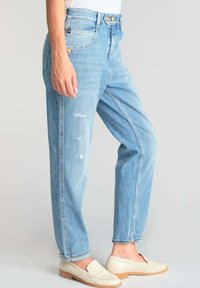 Jeans bleu clair taille haute avec une jambe droite, présentant des détails déchirés et une fermeture à boutons et à zip. Associés à des mocassins crème.