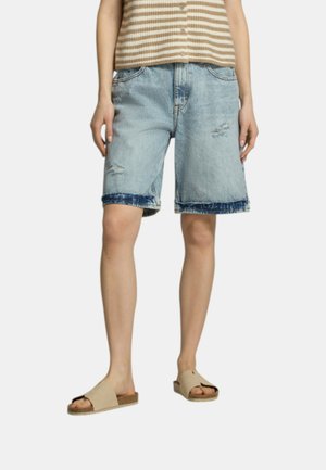 Donna che indossa pantaloncini in denim azzurro chiaro consumati, maglione smanicato a righe beige e sandali beige aperti sulle dita contro sfondo bianco.