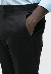 Δεξί χέρι εισερχόμενο στην μπροστινή τσέπη μαύρου παντελονιού που φοριέται με μανίκι ανοιχτό μπλε πουκάμισου.