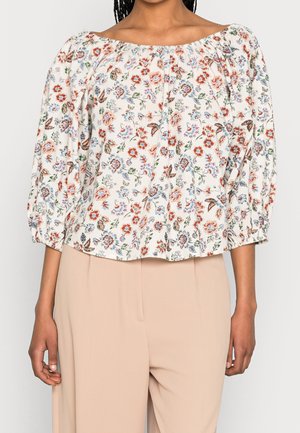 Mujer con blusa crema sin hombros con estampados florales en rojo, azul y verde y pantalones beige de cintura alta.
