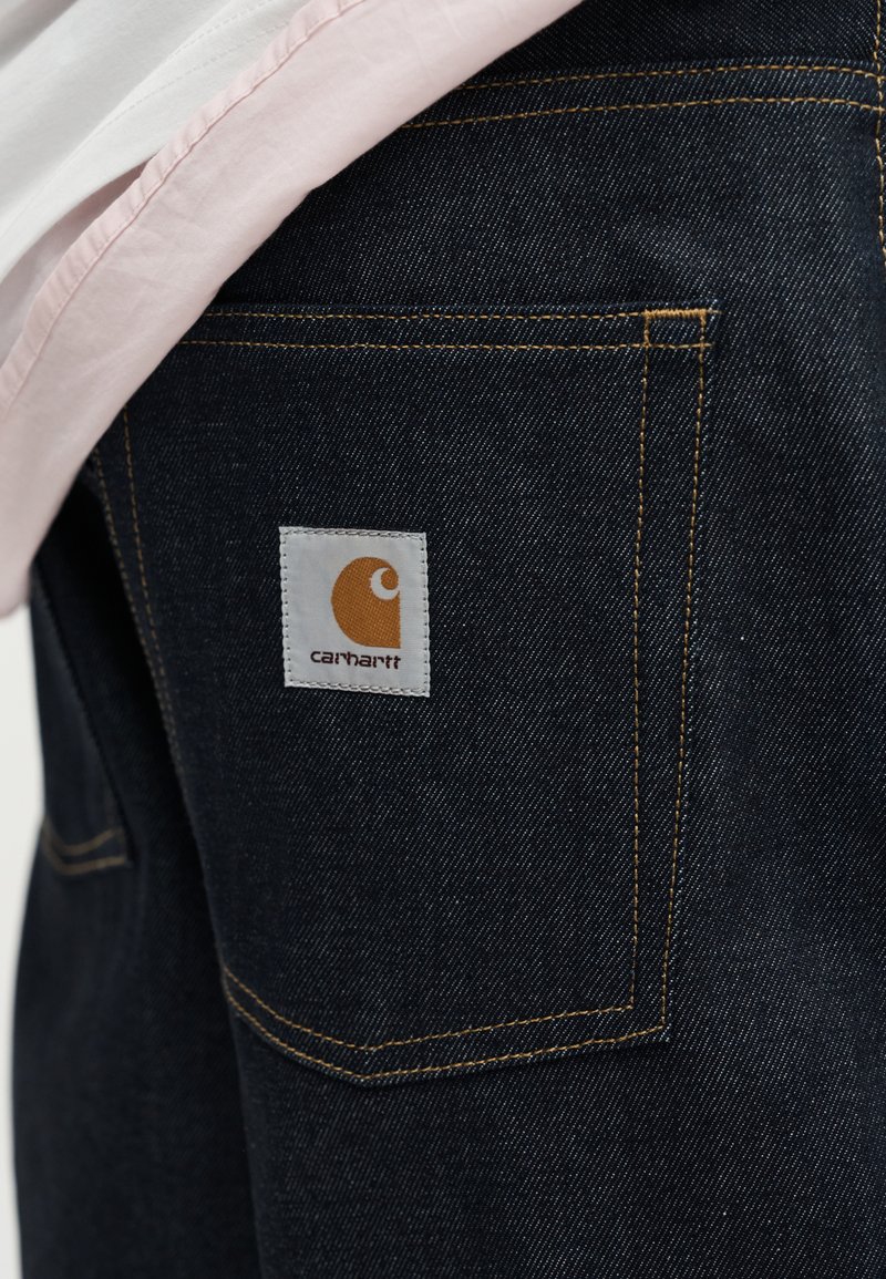 Tasca di jeans in denim blu scuro con cuciture gialle e un'etichetta quadrata bianca del marchio Carhartt, parzialmente coperta da tessuto rosa chiaro e bianco.