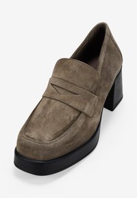 Mocasines de ante marrón con tacón, punta redondeada, costuras decorativas y tacón cuadrado. Suela de goma negra lisa para mayor tracción.