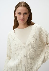 Crème kleurige gebreide cardigan met een V-hals, decoratieve zilverkleurige versieringen, gestructureerde patronen en drie knopen aan de voorkant.