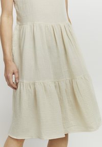 Helles beigefarbenes Kleid aus strukturiertem, gekräuseltem Stoff. Es hat einen taillierten Oberteil und einen ausgestellten, gestuften Rock. Keine sichtbaren Muster oder Akzente.