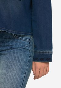 Giacca di jeans in tessuto blu scuro, con dettagli pattern sui polsini e un taglio dritto. Abbinata a jeans azzurri a righe verticali.