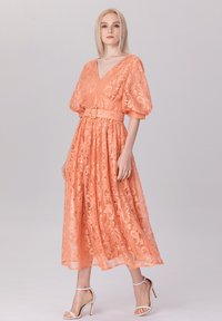 Robe longue en dentelle pêche avec un décolleté en V, des manches ballons et une taille ceinturée. Présente des motifs floraux et une jupe fluide.