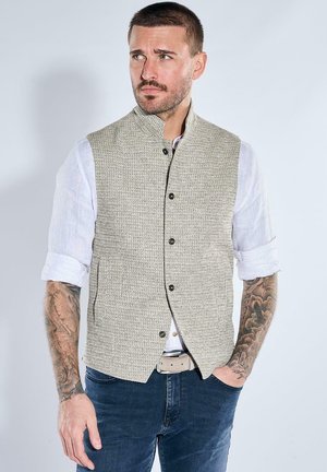 Man with short hair and tattoos wearing a buttoned beige textured vest over a white rolled-up shirt and dark jeans, against plain background.
