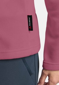 Pullover in pile rosa morbido con etichetta Polartec. Presenta una superficie liscia, una vestibilità sagomata e un orlo con tessuto blu a contrasto sotto.